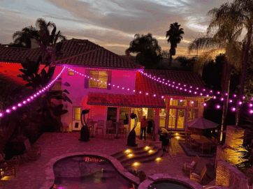 A backyard at dusk with a pool, patio furniture, and tropical plants. Purple string lights are draped across the area, illuminating the house and outdoor space with a warm, festive glow.