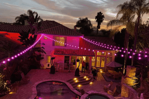 A backyard at dusk with a pool, patio furniture, and tropical plants. Purple string lights are draped across the area, illuminating the house and outdoor space with a warm, festive glow.