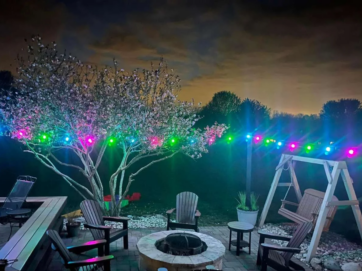 A backyard patio at night with a fire pit surrounded by chairs, a wooden swing, and a blooming tree illuminated by colorful string lights. The sky is dark with clouds and silhouetted trees in the background.