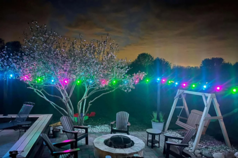 A backyard patio at night with a fire pit surrounded by chairs, a wooden swing, and a blooming tree illuminated by colorful string lights. The sky is dark with clouds and silhouetted trees in the background.