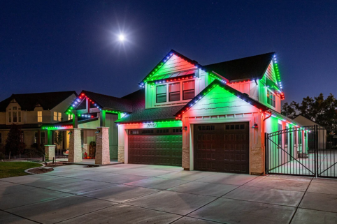 A two-story house at night is decorated with red, green, and white holiday lights along the roof. The sky is clear with a bright full moon shining above. The driveway and surrounding area are well-lit.