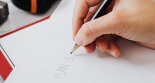 Attached image file A person sketches on white paper with a pencil at a desk, near a red notebook, a black tape measure, and a laptop.