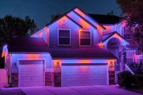 A two-story house at dusk is decorated with vibrant purple, pink, and blue LED lights outlining its roof, garage, and entryway, creating a colorful and festive appearance against the night sky.