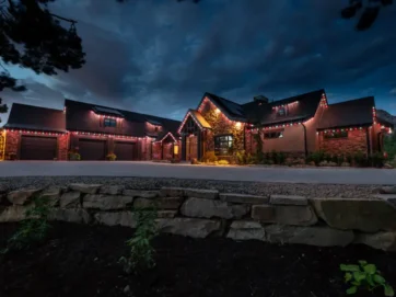 A large house with stone and wood exterior is illuminated by red and white string lights along the roofline at dusk, with a dark cloudy sky overhead and a stone wall and plants in the foreground.