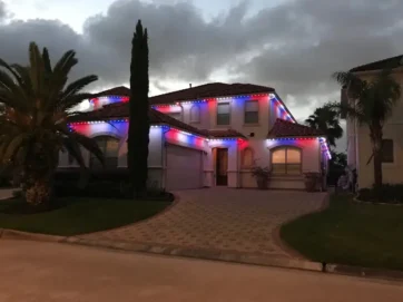 A two-story house is decorated with red, white, and blue lights along the roofline. Palm trees and shrubs are in the front yard, and the sky is cloudy at dusk.