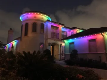 A two-story house is decorated with multi-colored string lights along the roofline, glowing in shades of red, green, blue, and purple at night. Cloudy sky and plants are visible in the foreground.